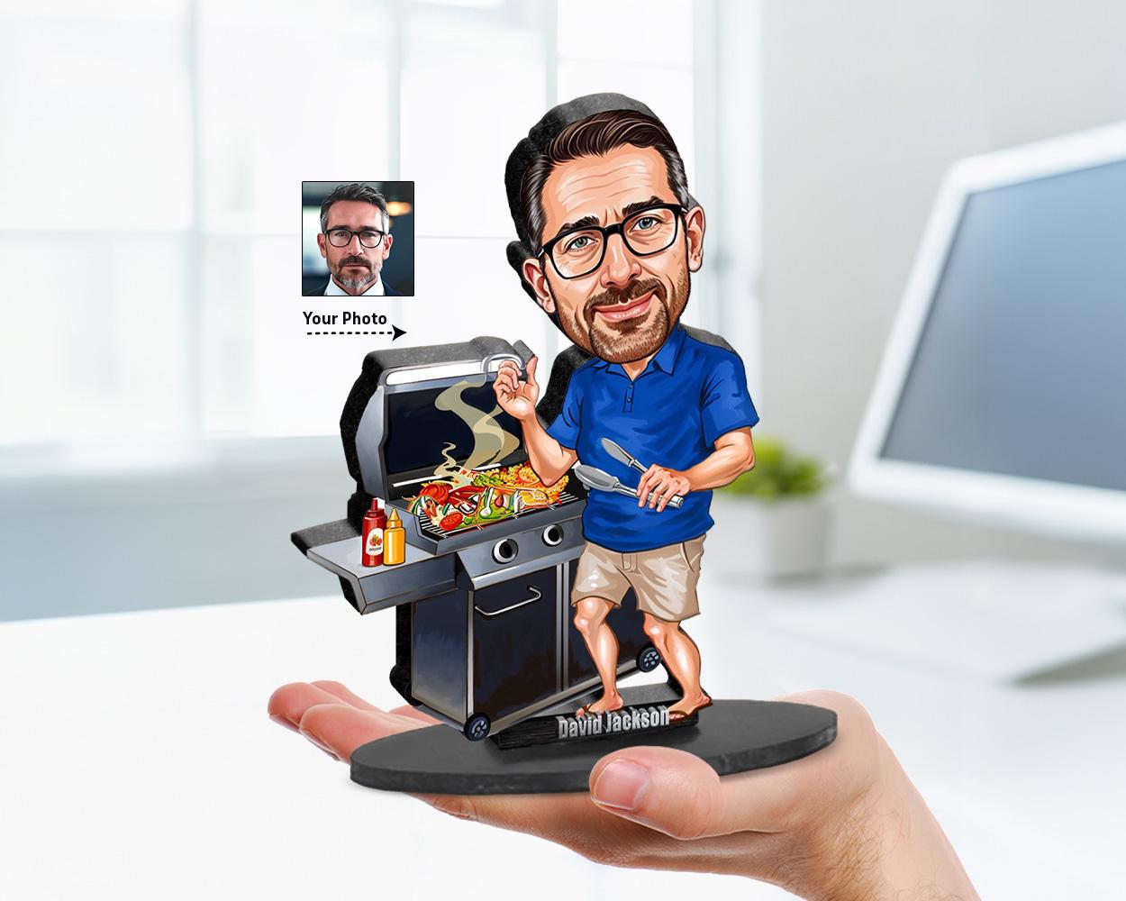 Big-head caricature of a male grill master standing at a BBQ grill, holding tongs and a fork. The figure is printed on wood or clear acrylic in a standing pose against a white background.